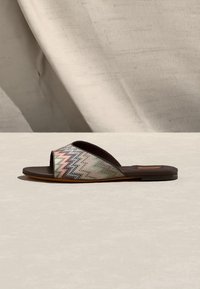 Σανδάλι με επίπεδη σόλα, ανοιχτή μύτη και επάνω μέρος από ύφασμα με πολύχρωμο ζιγκ-ζαγκ μοτίβο, σε ουδέτερη επιφάνεια με μπεζ φόντο.