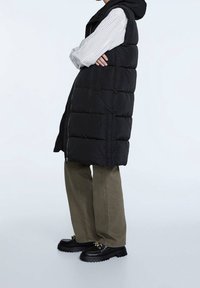 Zwarte puffer bodywarmer met capuchon, voorzien van een ritssluiting aan de voorkant, een doorgestikt ontwerp en twee zijzakken, gedragen over een gestreept shirt en olijfgroene broek.