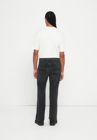 Άντρας με πλεγμένα μαλλιά που φοράει ένα απλό λευκό μπλουζάκι, μαύρα τζιν παντελόνια και μαύρα παπούτσια, στέκεται με άνεση μπροστά σε λευκό τοίχο.