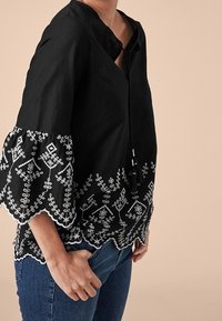 Blouse noire avec broderie florale blanche à l'ourlet et aux manches. Présente une coupe ample, un col en V et un détail à cordon.