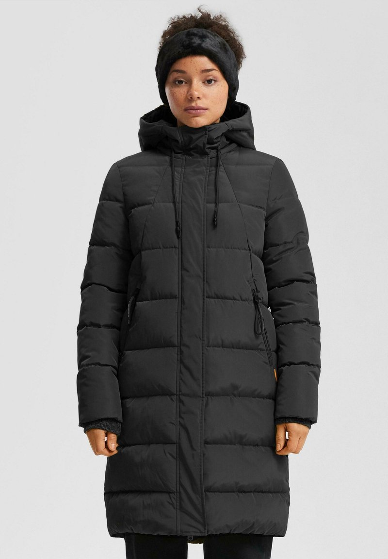 Sieviete, kas valkā garu melnu pūkaino mētelis ar kapuci un melnu pūkainu galvassegu, stāv uz vienkārša gaismas fona.