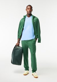 Survêtement vert avec des accents blancs, polo bleu clair et sac à dos vert texturé. Modèle portant des chaussures légères avec des semelles marron.