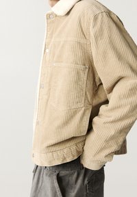 Beige kordfluweljacket med ett fårskinnskrage. Har synliga sömmar, två framfickor och en knäppning.