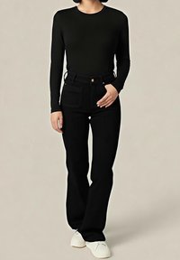 Personne portant un haut noir à manches longues et un pantalon noir taille haute avec des chaussures blanches, debout devant un fond clair uni.