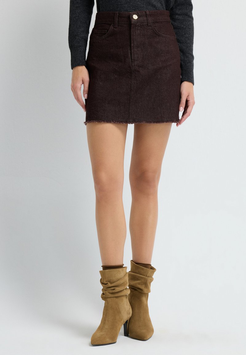 Vrouw in een donkerbruine corduroy mini-jurk, zwarte lange mouw top en tan suède slouchy haklaarzen.