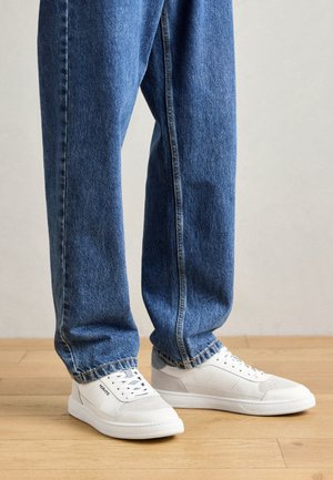 Valged tennisjalanõud kummist tallaga, külgedel perforatsiooniga detailid, väikesed heledad sinised teksad, puidust põrandal.