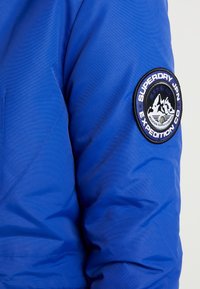 Veste isolante bleue avec un patch rond proéminent présentant des graphiques de montagne et du texte sur la manche droite. Tissu lisse et texturé.