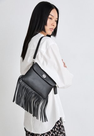 Vrouw met lang zwart haar, gekleed in een wit shirt en een rok met patroon, draagt een zwarte leren schoudertas met franjes en een zilveren sluiting.