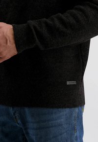 Donkere gebreide trui met een textuurafwerking, geribbelde manchetten en een klein merketiket aan de zijkant. Gemaakt in combinatie met blauwe denim jeans.
