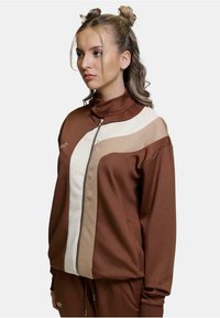 Brun zip-up jakke med høy krage, med kremfargede og beige aksentstriper. Laget av glatt, lett stoff.