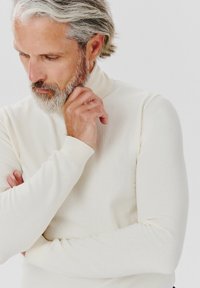 Sweter z golfem w kolorze kremowym, wykonany z miękkiego materiału, z dopasowanymi długimi rękawami i ściągaczami. Model ma siwe włosy i brodę.