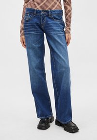 Μπλε τζιν παντελόνι με φαρδιά μπατζάκια από τζιν ύφασμα, μεσαίου ξεθωριάσματος, με αντίθετη ραφή και σχέδιο με πέντε τσέπες· συνδυασμένο με μαύρα παπούτσια πλατφόρμας.