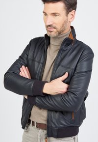 Zwarte leren bomberjack met ribbelmanchetten en -taille, heeft een ritssluiting, gestikte panelen en een bruine accent bij de kraag.