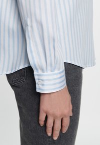 Ljusblå och vit randig blus med en mjuk textur, knappad manschett och en lätt rundad fåll, tillsammans med mörkgrå jeans.