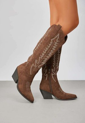Bottes en suede marron jusqu'au genou avec des bouts pointus, ornées de motifs brodés et fermées par une zip sur le côté. Talons épais et angulaires.