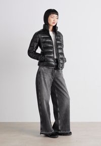 Svart boblejakke med et kviltet design, lange ermer, og glidelås foran, sammen med grå denimbukser med vid ben og svarte sko.