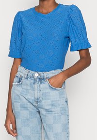 Top azul de manga corta con un patrón de ojales texturizados, que presenta mangas abullonadas y un cuello redondo. Combinado con jeans a cuadros en azul claro.