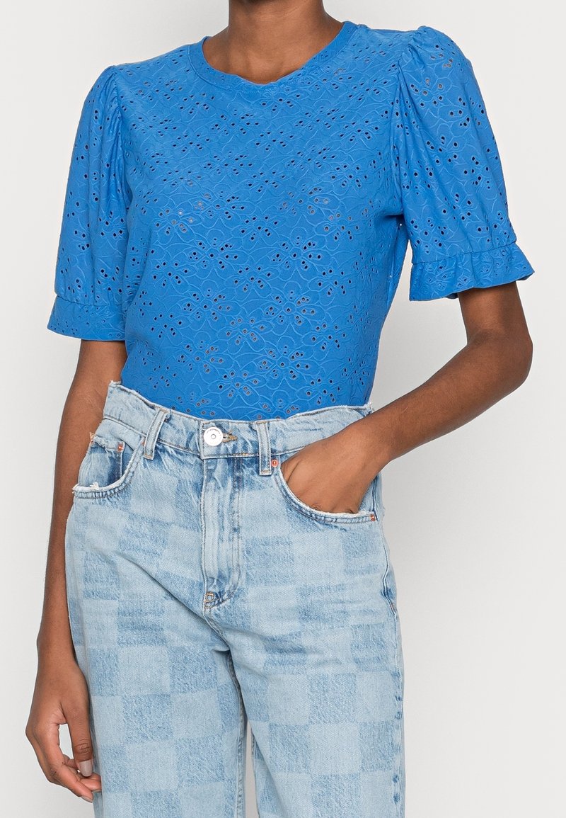 Top azul de manga corta con un patrón de ojales texturizados, que presenta mangas abullonadas y un cuello redondo. Combinado con jeans a cuadros en azul claro.