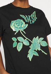 Camiseta de algodón negra con un gran diseño de una rosa metálica verde y hojas detalladas, y el nombre de la marca "DIESEL" arriba en una fuente en negrita.