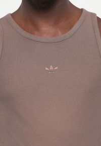 Brun, ribbet tanktop med rund halsudskæring; har et lille pink broderet logo på brystet. Stoffet ser blødt og tætsiddende ud.