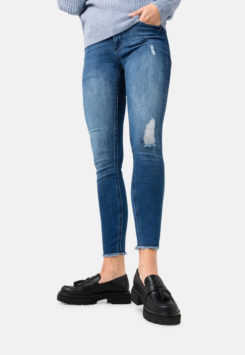 Jeans skinny in denim blu medio con dettagli strappati al ginocchio. Orlo sfrangiato abbinato a mocassini neri su uno sfondo bianco.