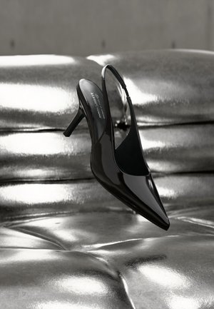 Czarny, lakierowany but na wysokim obcasie typu slingback, umieszczony na tle błyszczącej, metalicznej, srebrnej poduszki ze skóry.