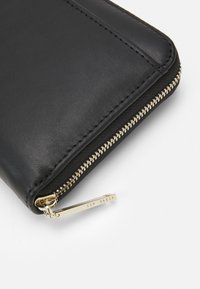Portafoglio in pelle nera con dettaglio della cerniera dorata. Presenta una texture liscia e un'etichetta metallica con il nome "Ted Baker" inciso.