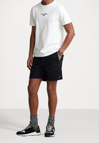 Man med vit Polo Sport t-shirt, svarta shorts med röd logga, svart och vit sneakers, gråspräckliga strumpor och armbandsur, stående med ena handen i fickan.