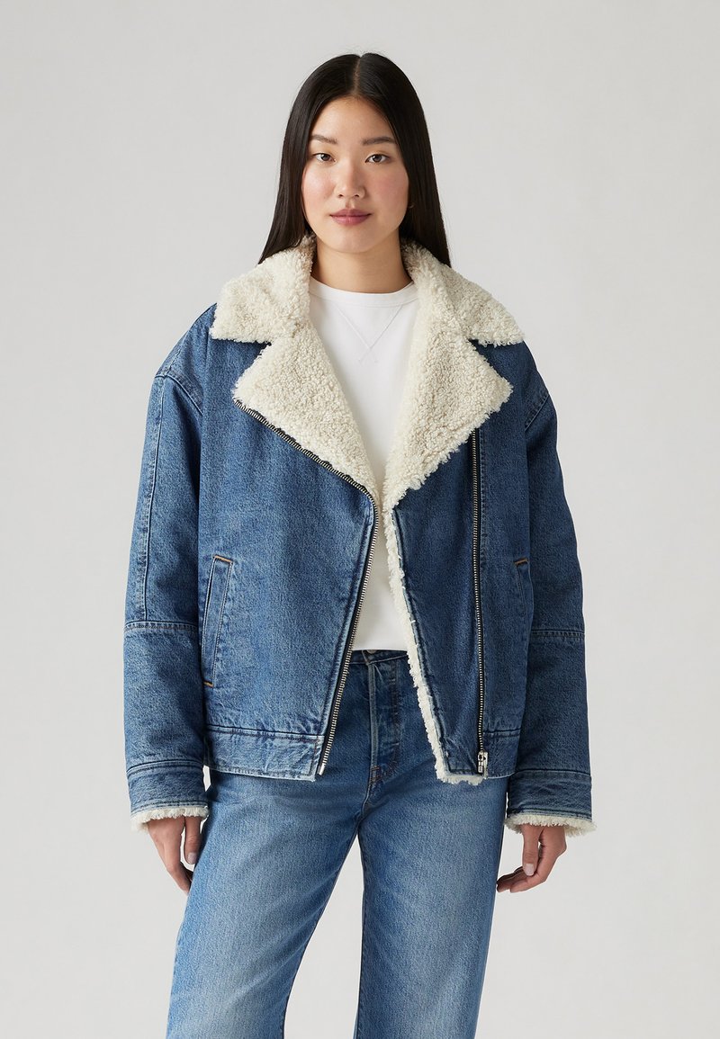 Veste en denim avec un col en fausse fourrure crème, coupe oversize, fermeture éclair, et poches latérales ; portée sur un top blanc et un jean bleu.