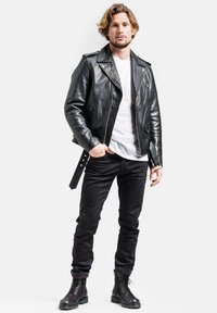 Schwarze Leder-Bikerjacke mit silbernen Reißverschlüssen, weißem T-Shirt darunter, schwarzen Jeans und schwarzen Schnürstiefeln. Das Modell steht vor einem weißen Hintergrund.
