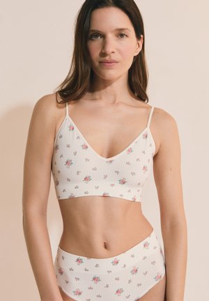 Woman wearing white floral ribbed bralette and matching high-waist briefs, standing against beige background, facing forward.