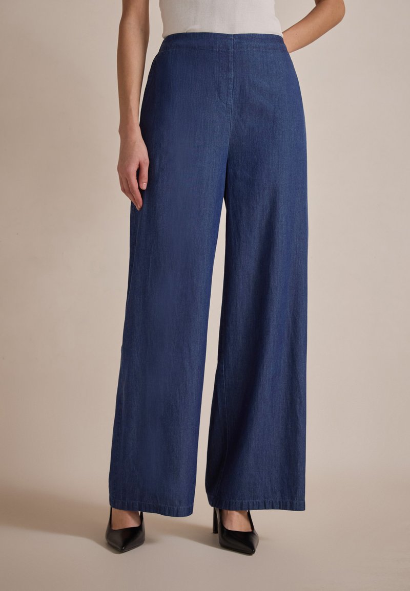 Pantalones de mezclilla oscuros de pierna ancha con una cintura alta, que presentan un diseño limpio, una textura sutil y bolsillos laterales, combinados con tacones negros.