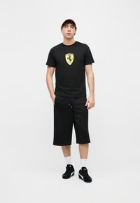 Νεαρός άνδρας đứng mặc áo thun đen với logo Ferrari, quần đùi dài màu đen, tất trắng, giày thể thao đen Puma và mũ trắng.