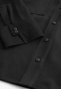 Černá košile s ribkovanou texturou, s knoflíkovými rukávy a sladěnými knoflíky na přední straně. Výrazné stehy podél okrajů.