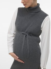 Kobieta w ciąży ubrana w szary prążkowany, bezrękawnikowy sweter, nałożony na białą bluzkę z długim rękawem, delikatnie trzyma brzuch jedną ręką.