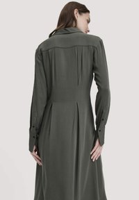 Eine Frau mit braunen Haaren trägt einen langen, dunkelgrünen, gefalteten Mantel mit geknöpften Ärmeln, von hinten vor einem schlichten, hellen Hintergrund betrachtet.