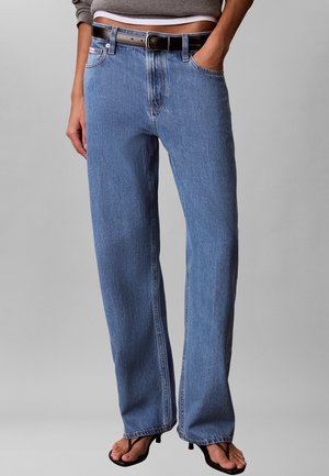 Person iført blå jeans med lige ben, sort bælte, grå cropped top og sorte åbne hæle-sandaler, stående med den ene hånd i lommen.