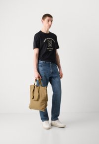 Sort t-shirt med korte ærmer og guld logo, blå denim jeans, lys beige tote bag med håndtag og hvide sneakers.