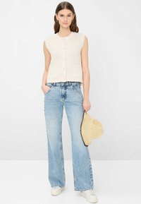 Donna che indossa un gilet beige senza maniche con bottoni, jeans larghi color azzurro chiaro, scarpe da ginnastica bianche, tiene un cappello di paglia nella mano destra.