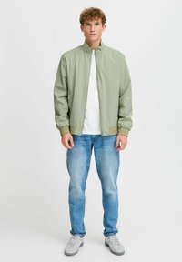 Lys grøn lett bomberjakke med ståkrage, elastiske mansjetter og nederkant. Båret over en hvit skjorte og kombinert med blå jeans.