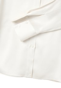 Camicia bianca a maniche lunghe con polsini abbottonati e plissettatura frontale, appoggiata su uno sfondo bianco.