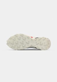 Semelle de chaussure de trail running avec un fond en caoutchouc blanc texturé, présentant des motifs de grip multidirectionnels et un accent rouge.
