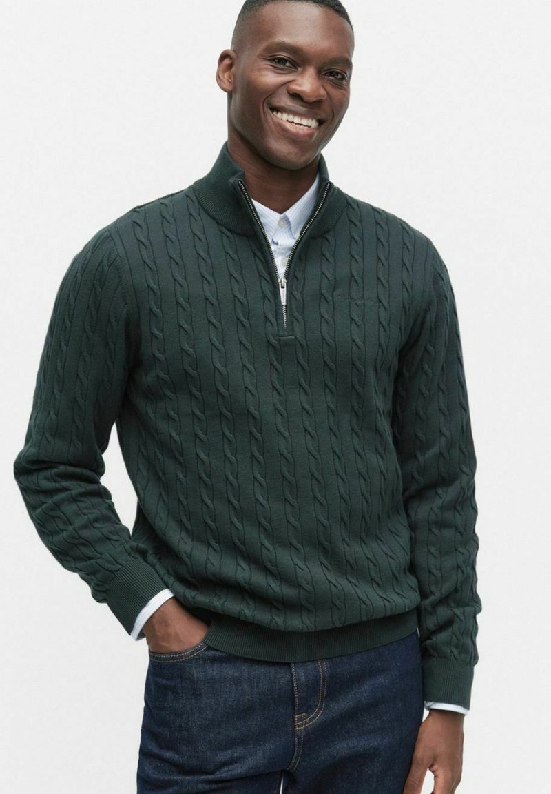 Mörkgrön kabelstickad tröja med kvartdragkedja i kragen, ribbade manschetter och nederkant, lagd över en ljusblå skjorta med krage.