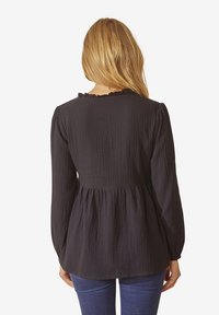 Eine Frau mit langen blonden Haaren, die eine schwarze Langarm-Bluse mit Struktur und blaue Jeans trägt, von hinten vor einem weißen Hintergrund gezeigt.