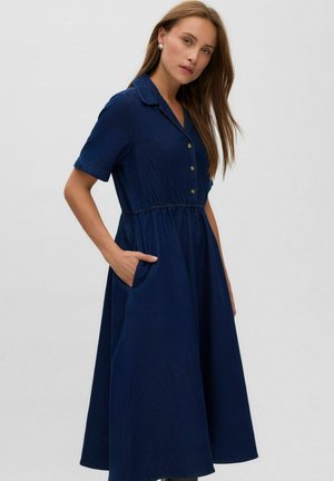 Femme portant une robe bleu marine à manches courtes boutonnée avec col, taille élastique et poches latérales, debout devant un fond uni.