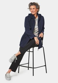 Marineblaues Kapuzenjackett mit einem schlanken Design, das über einen Knopfverschluss und zwei seitliche Taschen verfügt, kombiniert mit einem gestreiften Hemd und schwarzen Hosen. Graue Sneakers.