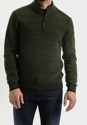 Homme portant un pull en maille côtelée vert foncé avec un col demi-zip et un jean bleu foncé, debout devant un fond uni.