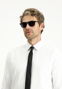 Occhiali da sole neri, camicia bianca con colletto e cravatta nera con trama. La camicia è aderente, con un aspetto di tessuto liscio.