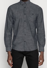Donkergrijs overhemd met knoopsluiting van een gestructureerde stof, lange mouwen en een enkele borstzak. Zwarte knopen contrasteren met de kleur van het shirt.