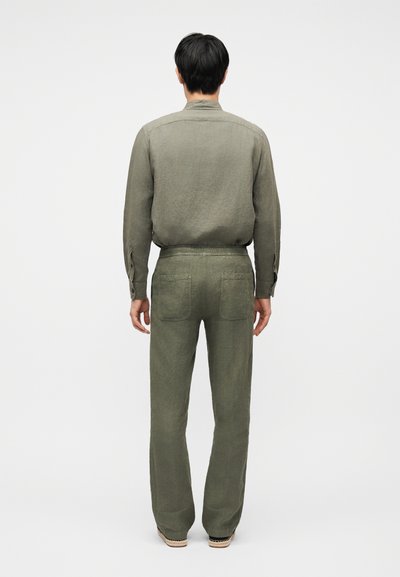 Άνδρας με την πλάτη να βλέπει, φοράει ομοιόμορφη μακρυμάνικη πράσινη ελιά πουκάμισο και παντελόνι με ίσια γραμμή και πίσω τσέπες, στέκεται σε απλό λευκό φόντο.